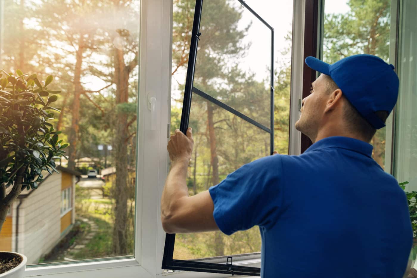 A man in a blue hat cleaning a window screen with solar screens. worker installing mosquito net wire screen on house window