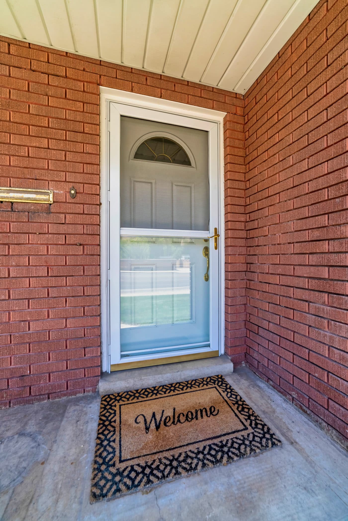 A welcome mat on the front door of a house. Gray front door and glass storm door at the entrance of home with red brick wall. Welcome doormat is also placed at the facade by the doorstep.