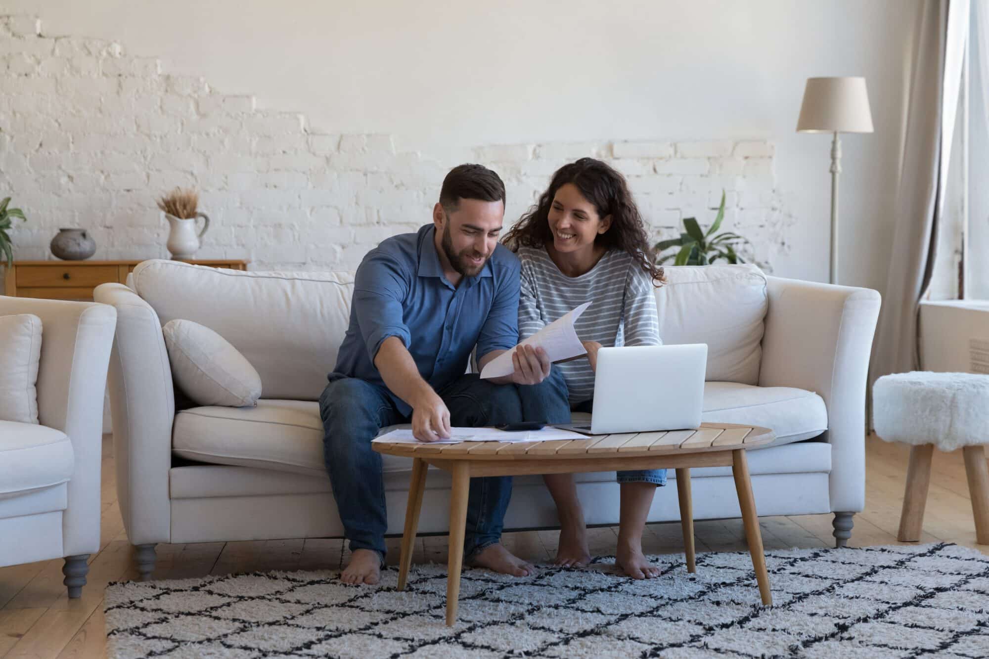 Contact couple on franchise site: Millennial wife and husband sorting out bank papers, check invoices, paying bills via e-bank on laptop sit on sofa smile feel satisfied. Enough money, payments, profit, financial management concept